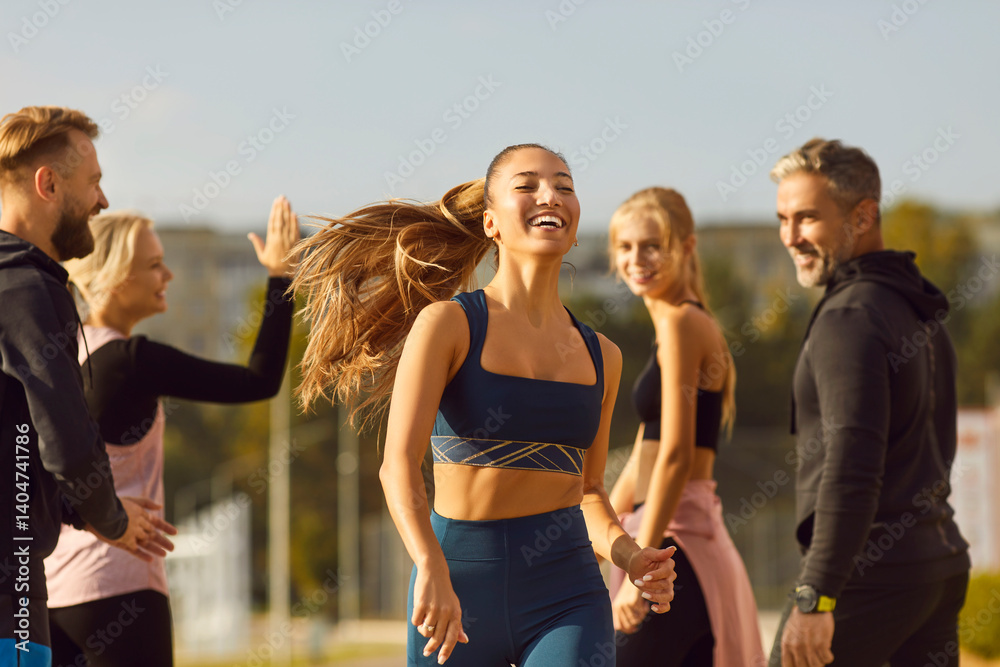 Portrait of young athletic girl running to the finish to friends supporting her standing in city park outdoors. Sporty woman doing jogging exercises in nature. Team sport and training concept.