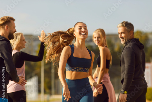 Obraz na płótnie Portrait of young athletic girl running to the finish to friends supporting her standing in city park outdoors. Sporty woman doing jogging exercises in nature. Team sport and training concept. w salonie