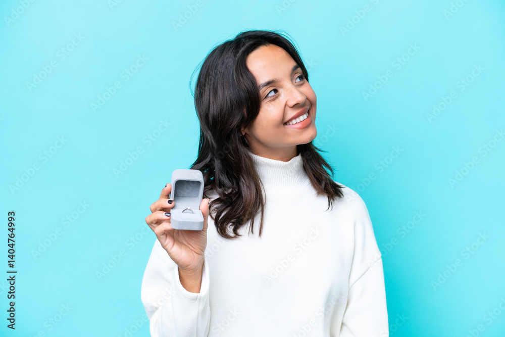 Young hispanic woman holding a engagement ring isolated on blue background thinking an idea while looking up Young hispanic woman holding a engagement ring isolated on blue background thinking an idea while looking up