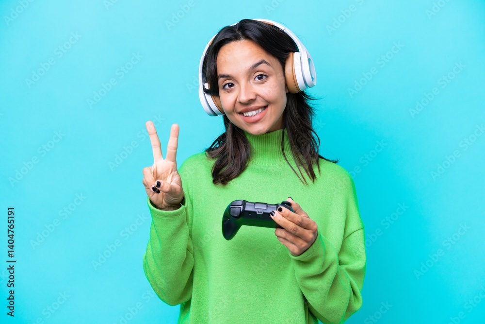 Young hispanic woman playing with a video game controller isolated on blue background smiling and showing victory sign Young hispanic woman playing with a video game controller isolated on blue background smiling and showing victory sign