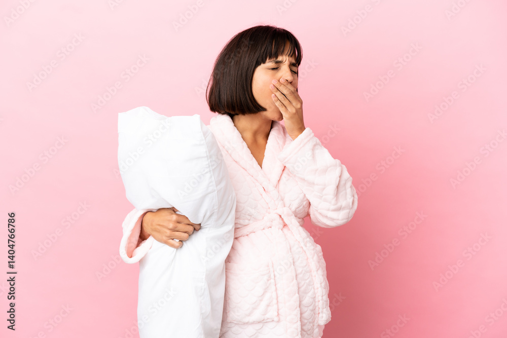Young mixed race pregnant woman isolated on pink background in pajamas and holding a pillow and yawning Young mixed race pregnant woman isolated on pink background in pajamas and holding a pillow and yawning