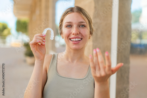 Young blonde woman holding invisible braces at outdoors inviting to come with hand. Happy that you came