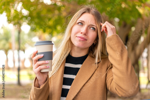 Young pretty blonde woman holding a take away coffee at outdoors having doubts and with confuse face expression