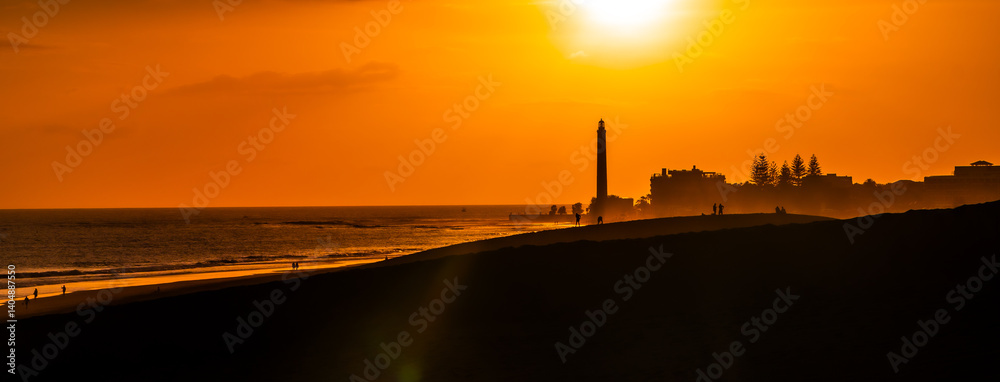 Tourists walking on maspalomas dunes at sunset with lighthouse in background