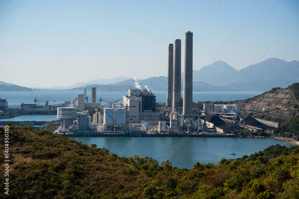 Naklejka premium Industrial power plant with tall chimneys on the waterfront of Lamma Island, Hong Kong, surrounded by green hills and overlooking a calm bay with mountains in the background