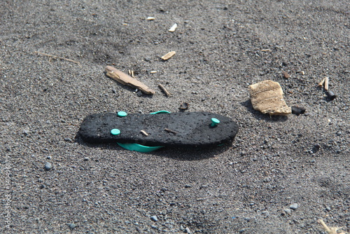 A worn-out black flip-flop with teal straps lies abandoned on a sandy surface with scattered debris. Lost footwear