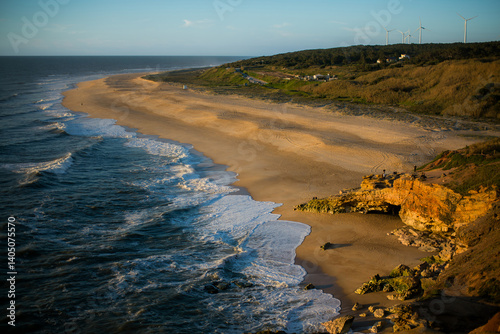 Obraz na płótnie A sunlit beach in Nazaré, Portugal, on a clear day, with golden sands meeting the serene Atlantic Ocean under a bright blue sky. w salonie