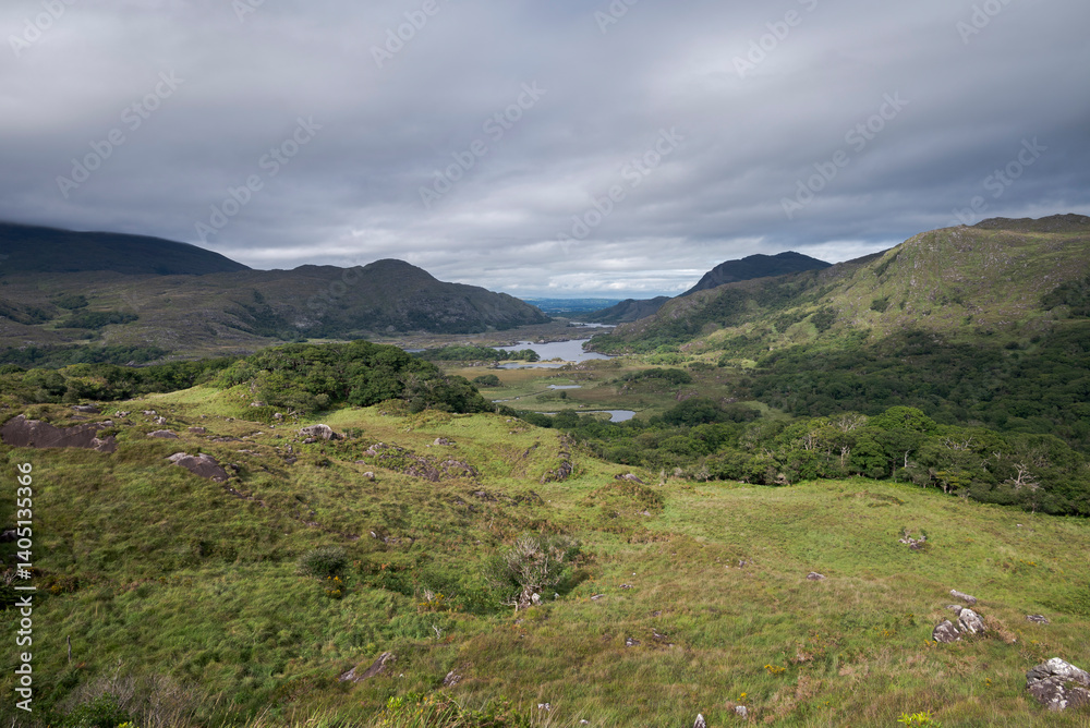 A scenic view of Derrycunnihy Wood in Ireland, showcasing lush greenery, rolling hills, and a serene body of water under a cloudy sky