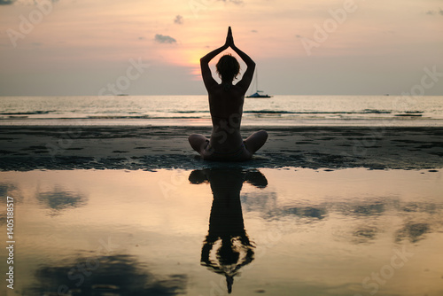 Obraz na płótnie The silhouette of a woman in a lotus pose, practicing yoga on a tropical beach at twilight in Thailand, her reflection shimmering in the calm water. w salonie