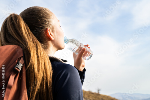 Thirsty hiker woman with backpack drinking water from a plastic bottle, standing in the mountains. Trekking, hiking, maintain balance of body preventing dehydration outdoors wellness healthy lifestyle