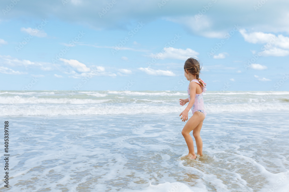 rear view of little girl in the swimsuit standing on the beach playing with waves rear view of little girl in the swimsuit standing on the beach playing with waves