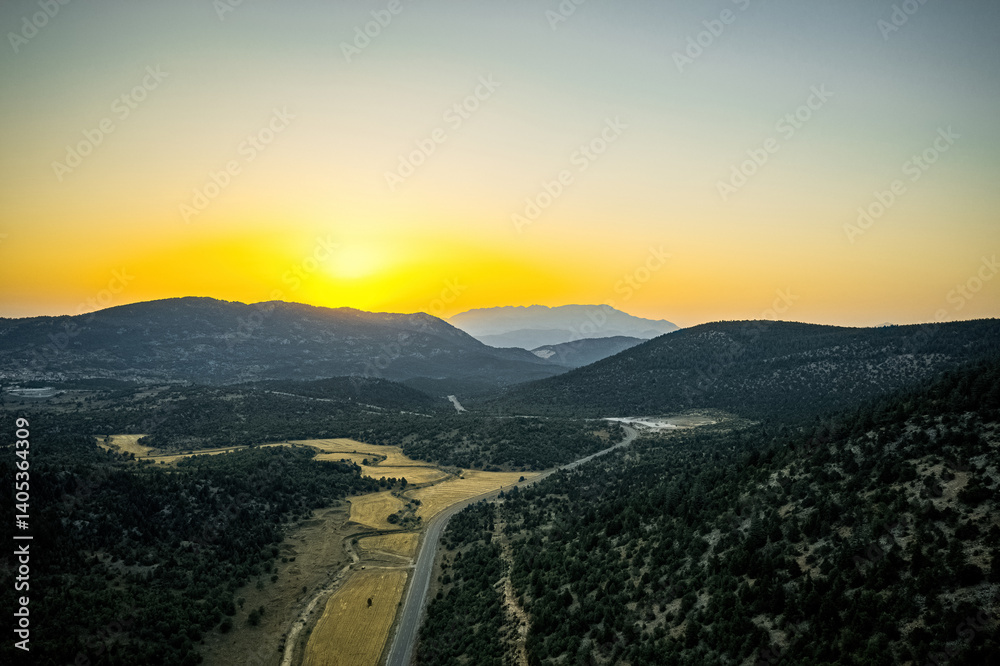 Winding asphalt mountain road with green forest area surrounded by mountainous trees on both sides in sunset landscape. Drone footage.