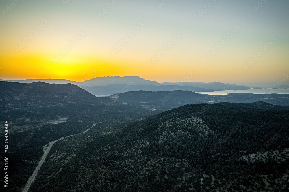 Winding asphalt mountain road with green forest area surrounded by mountainous trees on both sides in sunset landscape. Drone footage.