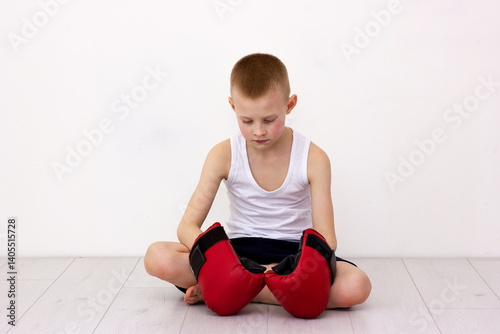 A boy sits on the floor wearing boxing gloves after a defeat. Sad look. Frustrated feelings, resentment and anger after failure