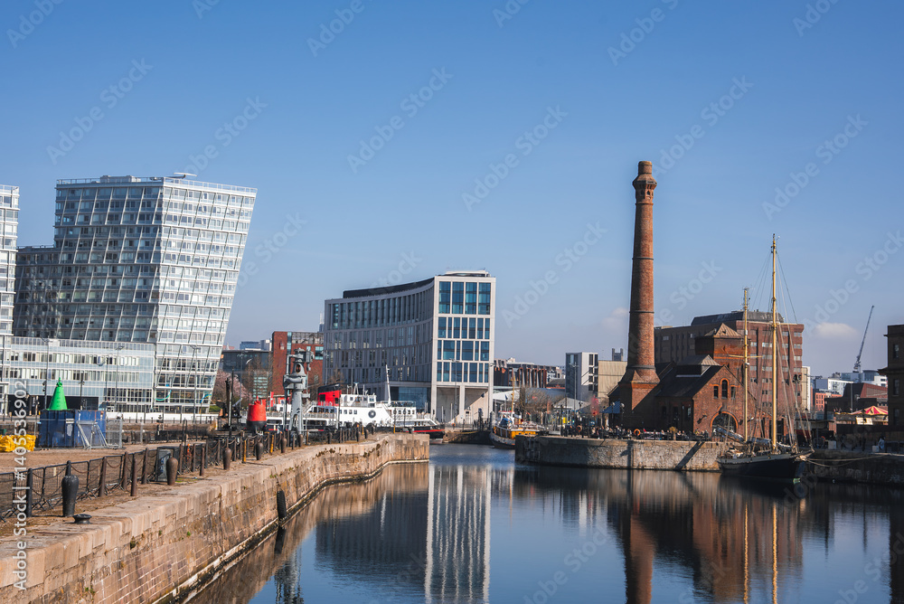 View of Liverpool's waterfront featuring Albert Dock, the Pump House chimney, and modern glass buildings reflected in calm water. View of Liverpool's waterfront featuring Albert Dock, the Pump House chimney, and modern glass buildings reflected in calm water.