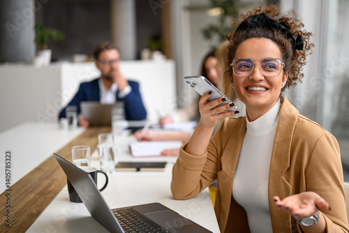 Female manager talking confidently on mobile phone's speaker with professional coworkers collaborating in the background