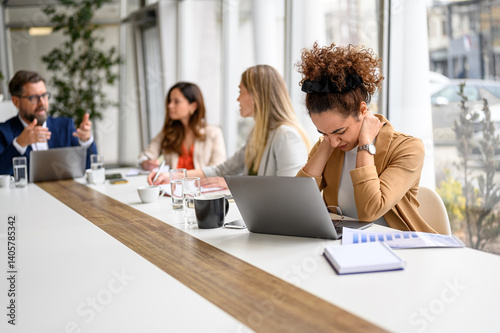 Stressed businesswoman holding her neck in pain and working on laptop while colleagues discussing in meeting