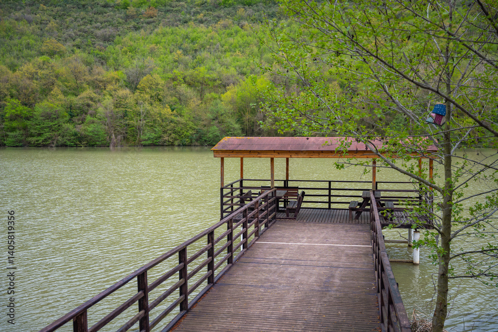 Obraz premium Wooden pier with gazebo over lake and forest backdrop. Serene lakeside nature spot. Rustic wooden canopy with red-roofed gazebo over calm lake waters and green hills. Relaxation concept