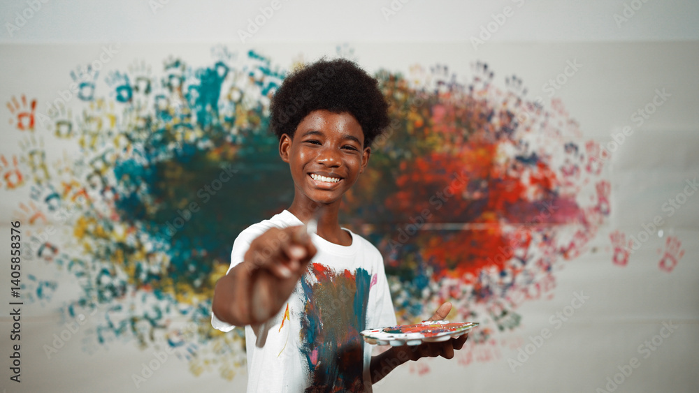 African boy standing at colorful stained wall while holding brush and point at camera. Happy highschool student wearing white shirt with stain color and look at camera with confident. Edification.