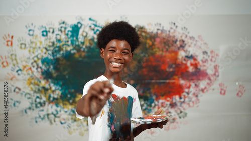 Obraz na płótnie African boy standing at colorful stained wall while holding brush and point at camera. Happy highschool student wearing white shirt with stain color and look at camera with confident. Edification. w salonie