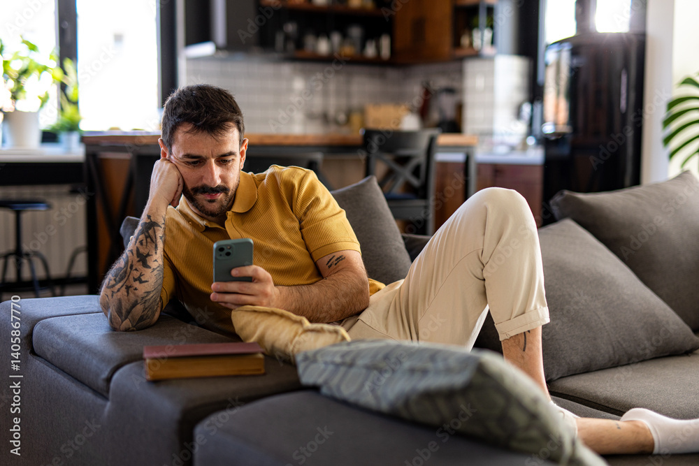 Obraz premium Relaxed young man lying on a comfortable sofa in a modern living room, using his mobile phone while enjoying his leisure time