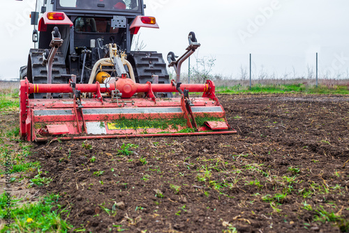 A tractor equipped with a red rotary cultivator actively prepares the soil in a rural field. The equipment loosens the earth, preparing it for planting and cultivation