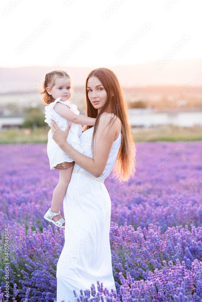 Naklejka premium Beautiful young mother holding baby girl 1 year old wearing white rustic dresses in blooming lavender field outdoors. Summer season.