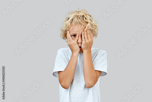Shy and curious, an 8 years old blonde boy with curly hair hides his face behind hands, peeking through fingers in a light t-shirt on grey background.