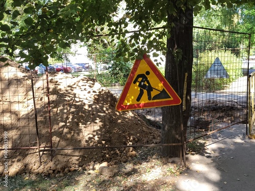Construction site on city street is marked by metal barricade with triangular warning sign depicting road work