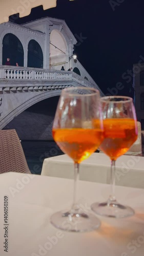 Two glasses of Aperol Spritz sit on a table with the Rialto Bridge in Venice, Italy, blurred in the background at night. A perfect Italian evening.