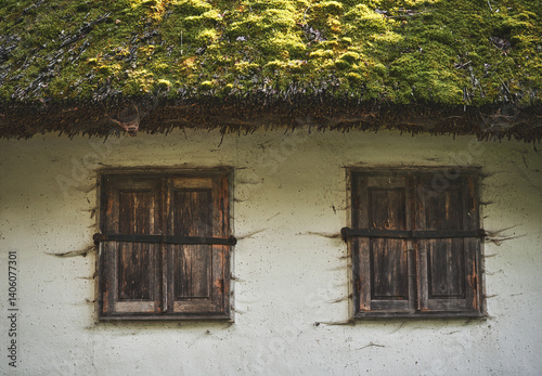 Part of an abandoned old traditional house with wooden windows and a slate roof covered moss and lichen. Two brown windows with closed shutters covered cobwebs on a white wall of a clay house
