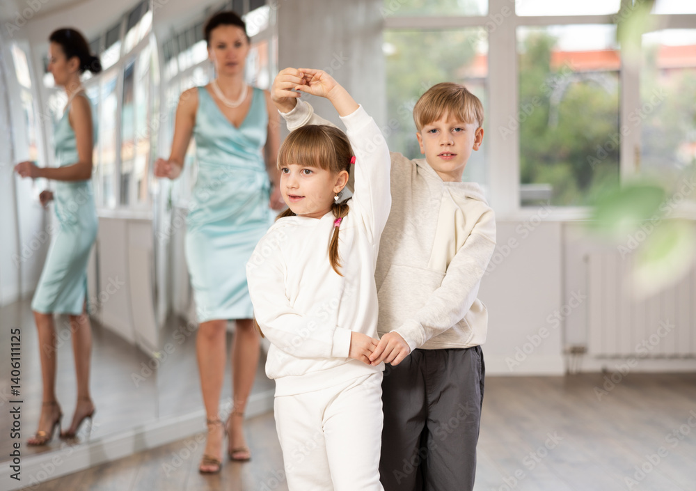 Motivated preteen ballroom dancers, girl and boy in sportswear practicing elegant dance moves in pair in bright studio, with female coach supervising in background.. Motivated preteen ballroom dancers, girl and boy in sportswear practicing elegant dance moves in pair in bright studio, with female coach supervising in background..