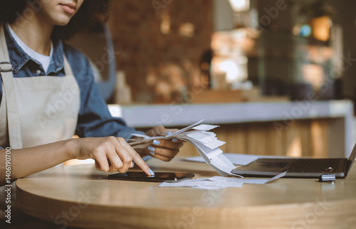 Reporting and accounting during covid-19 quarantine. Busy african american lady owner in apron works with financial bills and calculates profit and debt in modern cafe, empty space, cropped