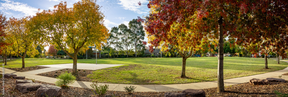 Fototapeta premium Panoramic view of a suburban park in Werribee, Melbourne, Australia with autumn foliage, a public basketball court, green open lawns, and pedestrian pathways.