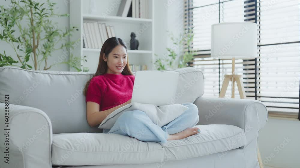 Happy young Asian woman using laptop while seated on couch at home Happy young Asian woman using laptop while seated on couch at home