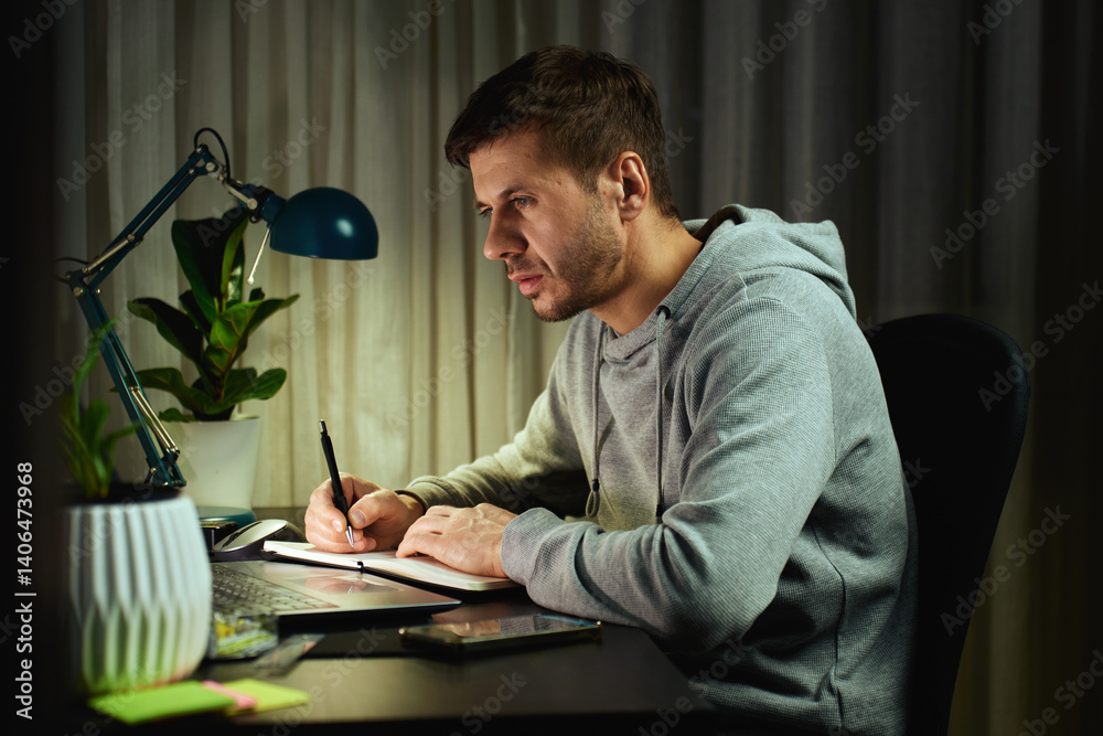 Concentrated freelancer working late at home office. Focused man in hoodie writes notes while working on laptop. Concept of night work