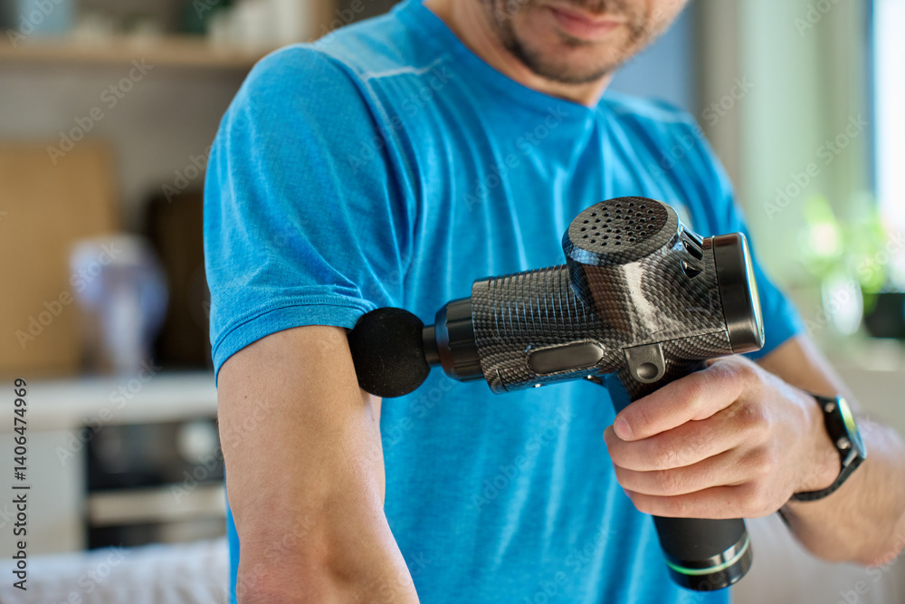 Athletic man using percussion massage device indoors. Man applies massage gun to arm muscle while sitting in living room, recovery therapy after exercise. Concept of home fitness and relaxation