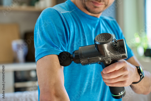 Obraz na płótnie Athletic man using percussion massage device indoors. Man applies massage gun to arm muscle while sitting in living room, recovery therapy after exercise. Concept of home fitness and relaxation w salonie