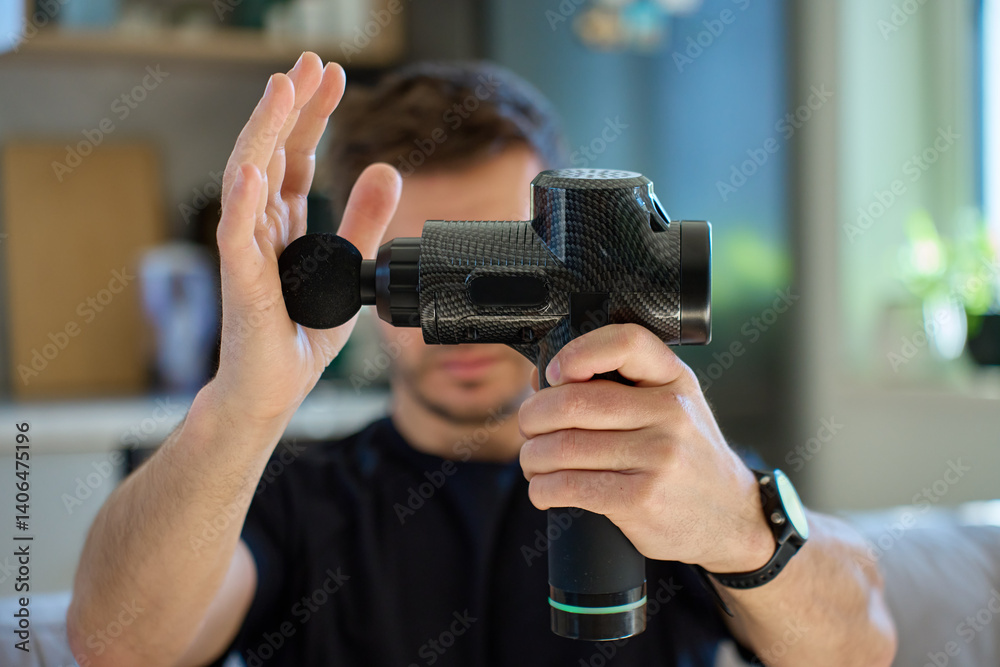 Close-up of man holding massage gun and pressing its vibrating head against his hand to test its strength and performance. Concept of sports recovery, self-care, and muscle treatment tools