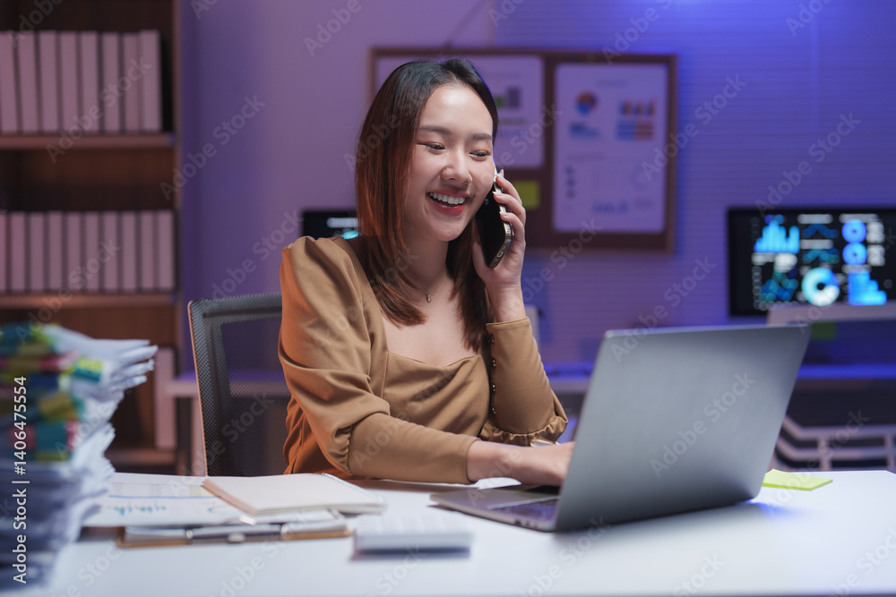 Young happy professional working overtime at night using laptop and speaking on mobile phone while sitting at desk in office with graphs and charts showing on screens Young happy professional working overtime at night using laptop and speaking on mobile phone while sitting at desk in office with graphs and charts showing on screens