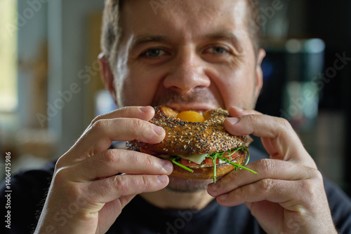 Obraz na płótnie Man takes big bite of seeded bagel filled with egg, cheese, tomato and greens. Hungry man eats sandwich at kitchen, close up w salonie