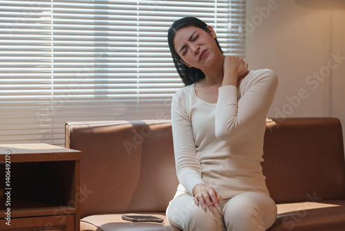 Young woman massaging her neck with a pained expression, experiencing discomfort from stiff muscles after prolonged hours of sedentary work at home on the sofa