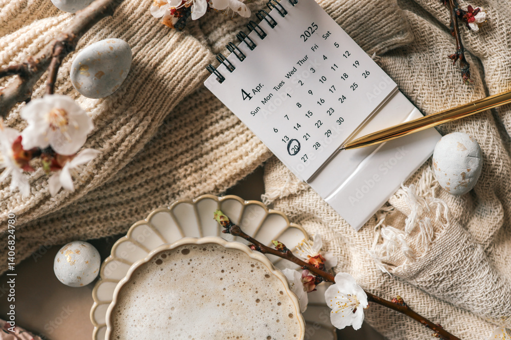 Fototapeta premium April 2025 calendar with April 20 marked, next to a golden pen, cup of coffee, blooming spring branches, and decorative Easter eggs on a knitted beige fabric background