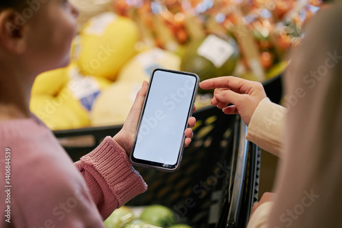 Person showing someone else smartphone in grocery store with several fruits and vegetables visible in background. Engaging in discussion about information displayed on screen