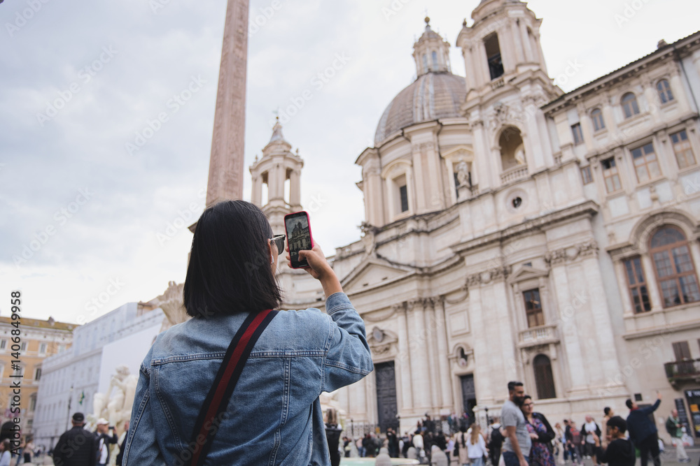 Fototapeta premium Young woman tourist is taking a photo of sant'agnese in agone and the obelisk using her smartphone in the famous piazza navona in rome, italy