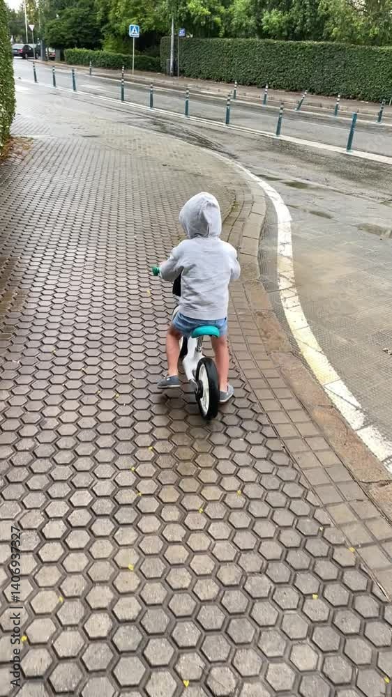 A young child wearing a gray hoodie rides a balance bike along a wet, tiled sidewalk after the rain. The scene captures a moment of independence, play, and exploration in an urban setting A young child wearing a gray hoodie rides a balance bike along a wet, tiled sidewalk after the rain. The scene captures a moment of independence, play, and exploration in an urban setting
