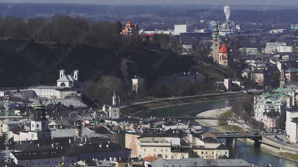 Aerial view of Salzburg, Austria, showing the Salzach River, St. Andra Church, historic buildings, moving vehicles, and distant industrial smoke.