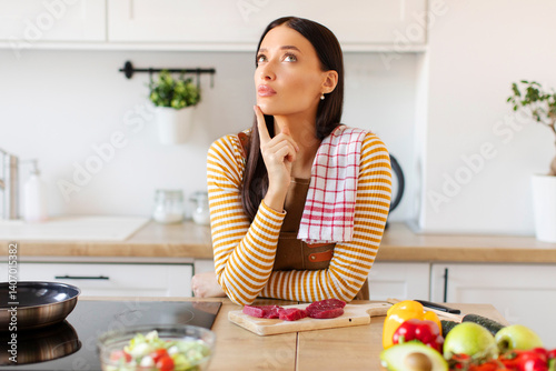 Photos Pensive woman touching chin, reminding recipe while cooking tasty dinner at home