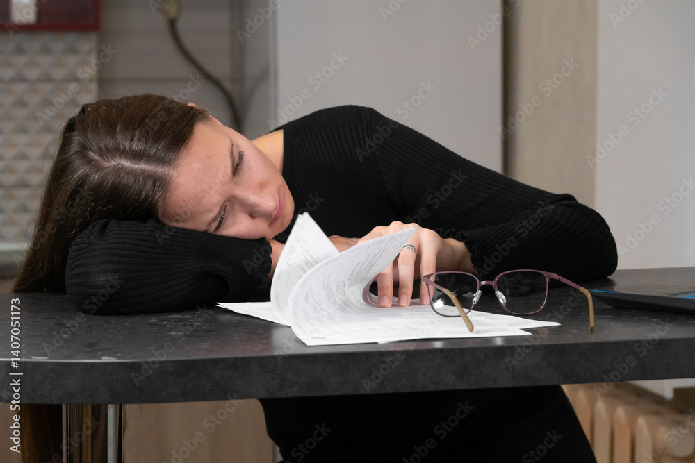Young woman falling asleep while working or studying from home, resting head on kitchen table with paperwork and eyeglasses, depicting exhaustion, stress, and burnout