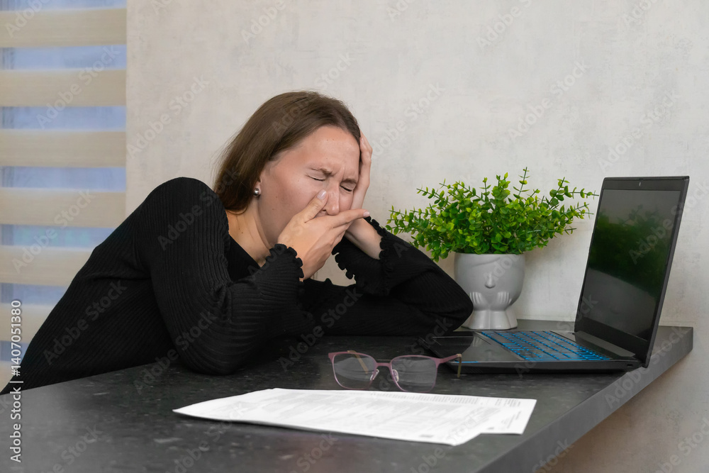 Young woman yawning and covering her mouth with her hand, feeling tired and exhausted while working from home in front of her laptop, struggling with fatigue and stress